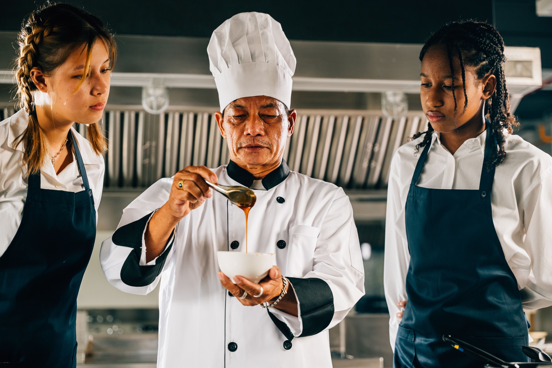Professional Asian senior chef instructs diverse students in a restaurant kitchen focusing on teamwork learning and note-taking. Educational workshop. Food Edocation